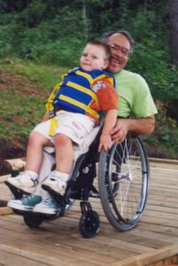 Older man in a wheelchair on a dock. He holds a young boy on his lap.