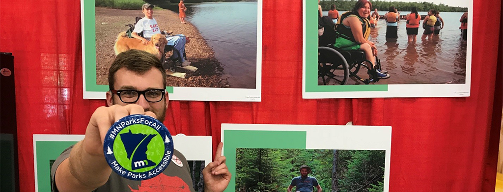 Man holding a button that reads: ”MN Parks For All, Make Parks Accessible.” In the background are photos of people with disabilities enjoying state parks.