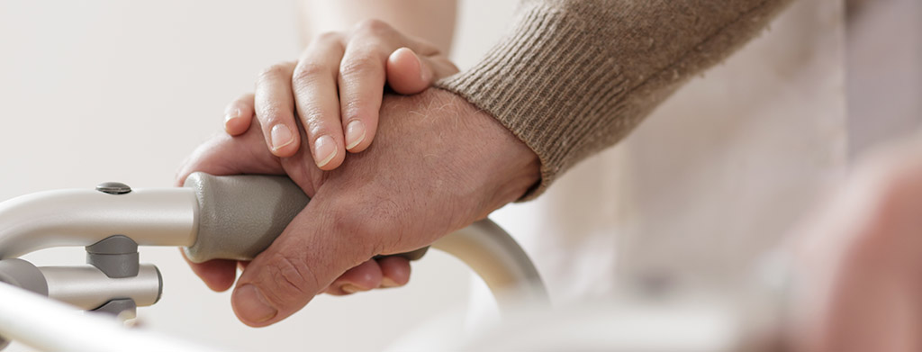 Close up of a man's hand holding on to a walker. A woman's hand rests on top of his.