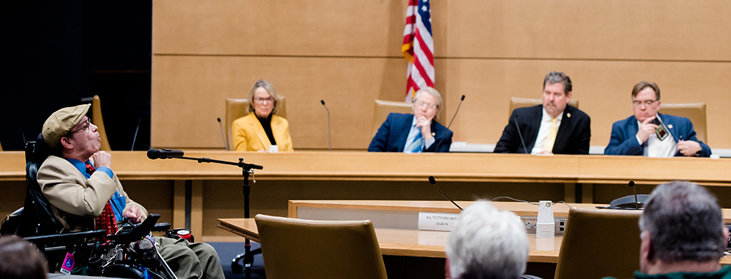 Man who uses a wheelchair speaking to four legislators, American flag in background