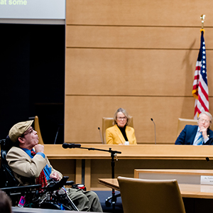 Man who uses a wheelchair speaking to legislators, American flag in background