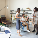 Three coworkers talking in an office. The coworker in the middle has an amputated right leg and is using a wheelchair.