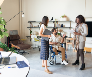 Three coworkers talking in an office. The coworker in the middle has an amputated right leg and is using a wheelchair.