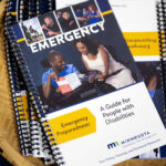A pile of emergency preparedness guides laying in a basket. The cover features photos of people with disabilities, including two people in wheelchairs looking at the guide together. Text reads: "Emergency Preparedness: A Guide for People with Disabilities. Minnesota Council on Disability. Your Policy, Training, and Technical Resource."