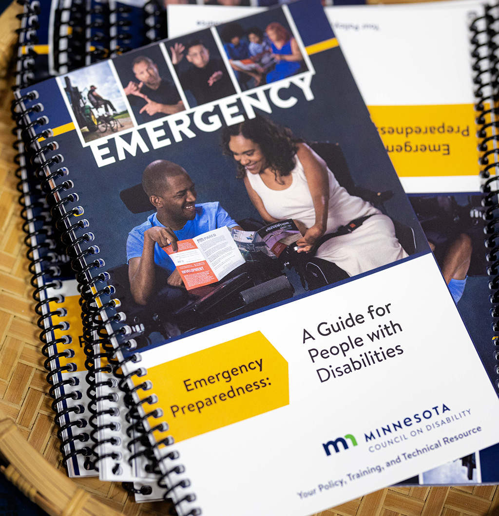 A pile of emergency preparedness guides laying in a basket. The cover features photos of people with disabilities, including two people in wheelchairs looking at the guide together. Text reads: "Emergency Preparedness: A Guide for People with Disabilities. Minnesota Council on Disability. Your Policy, Training, and Technical Resource." 