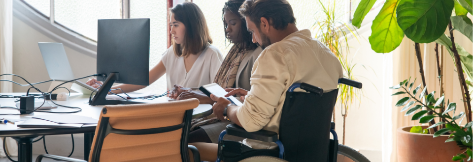 Three people working around a computer that sits on a table. One of them uses a wheelchair.