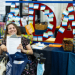 Woman who uses a wheelchair, holding a copy of the public policy survey and smiling. She is sitting in front of a table with a red ADA sign at the MCD State Fair booth.