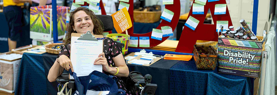 Woman who uses a wheelchair, holding a copy of the public policy survey and smiling. She is sitting in front of a table with a red ADA sign and a box of “Disability Pride!” posters at the MCD State Fair booth.