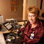 Woman using a wheelchair sits at her desk. In front of her are several electronic devices, a modified keyboard, and a telephone.