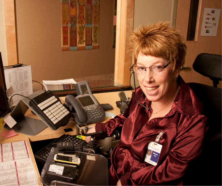 Woman using a wheelchair sits at her desk. In front of her are several electronic devices, a modified keyboard, and a telephone.