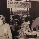 Black and white photo. Three women working behind a table. A sign above them reads, "Governor's Conference on the Handicapped."