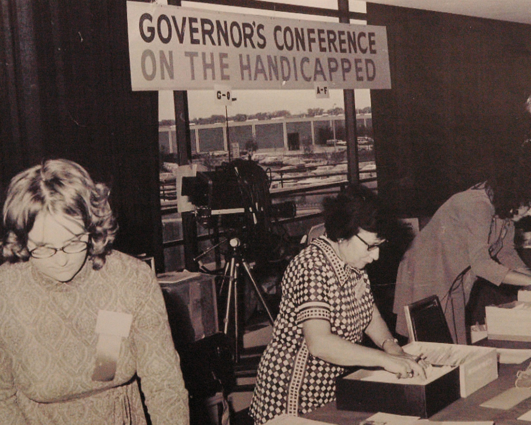 Black and white photo. Three women working behind a table. A sign above them reads, "Governor's Conference on the Handicapped."