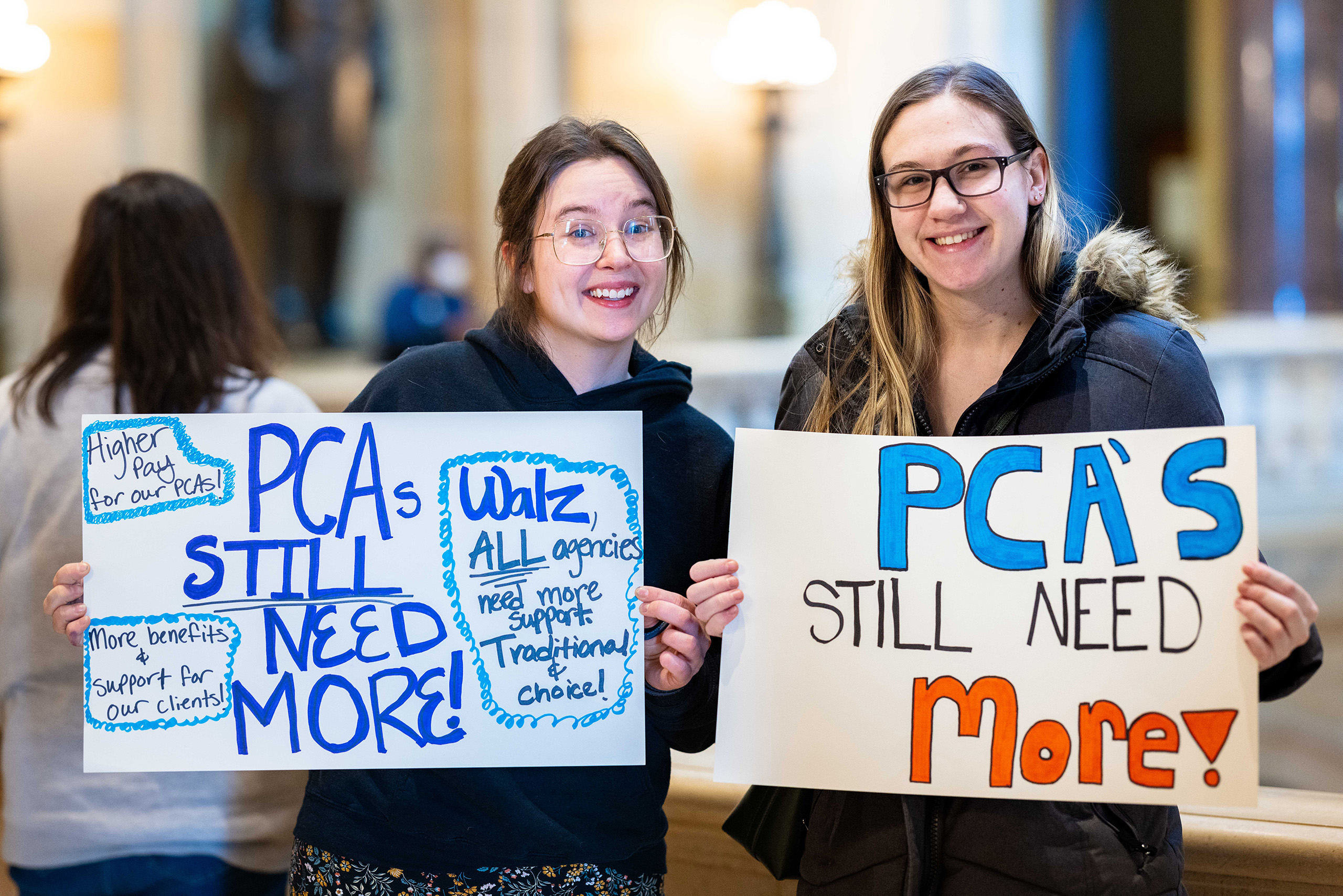 Two young women holding signs that read, "PCAs Still Need More!"