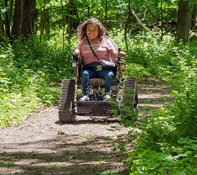 A woman using an all-terrain electric track chair on a wooded trail.
