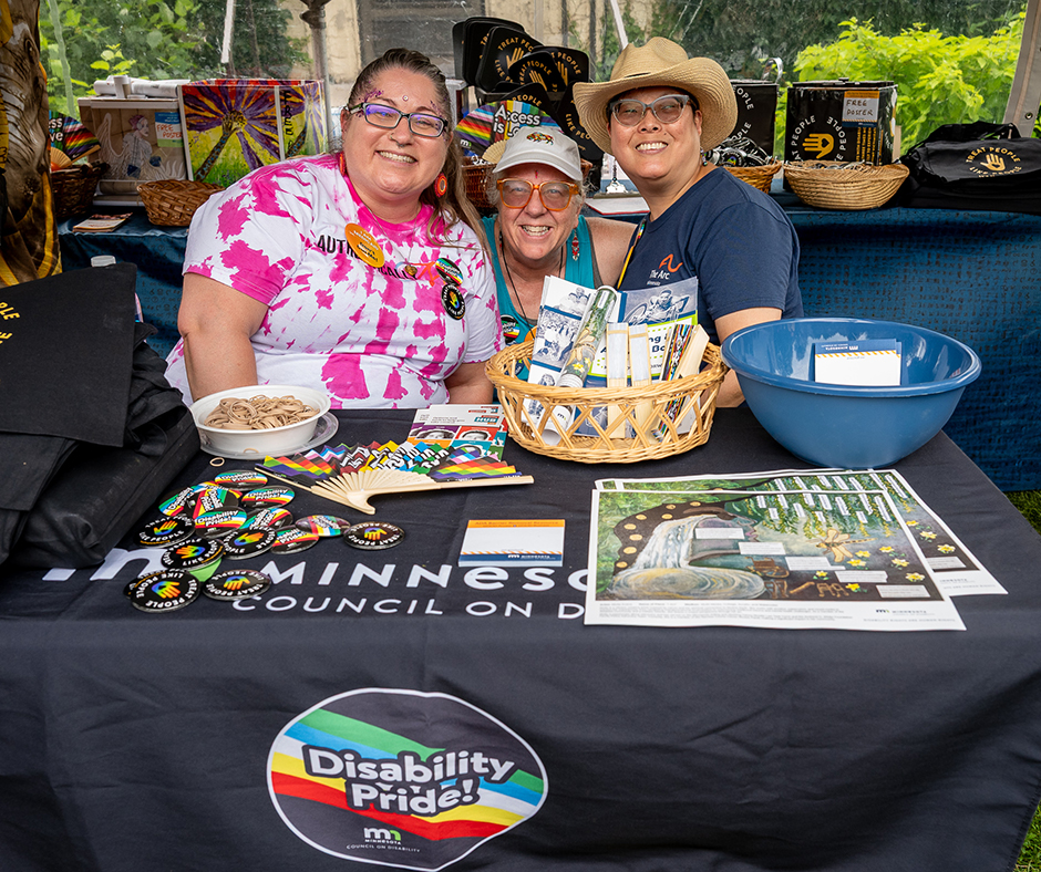 Three people smile behind the MCD booth at Twin Cities Pride, with Pride-themed materials and giveaways on the table.