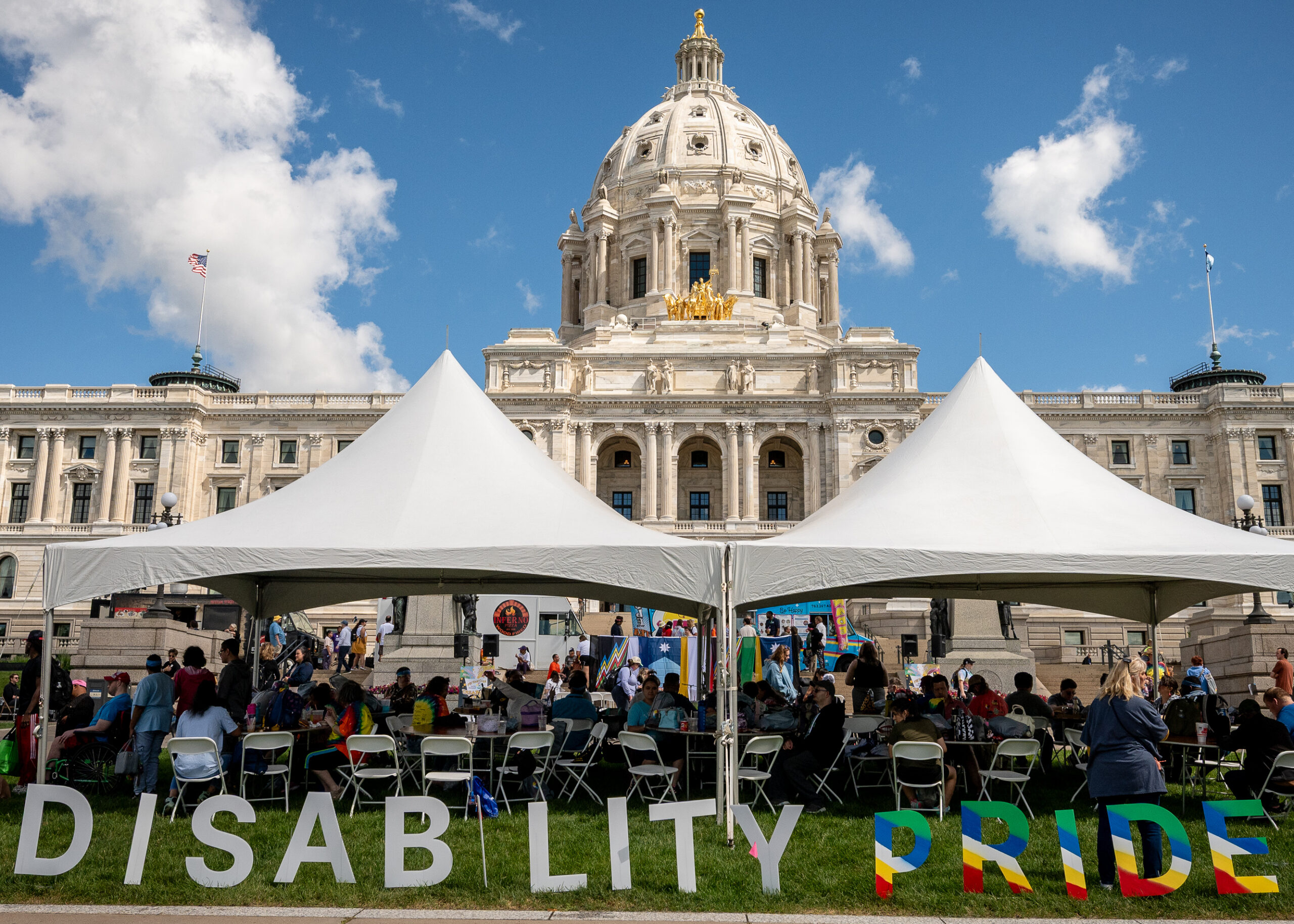 People gather under tents outside the Minnesota State Capitol behind large letters spelling “Disability Pride.”